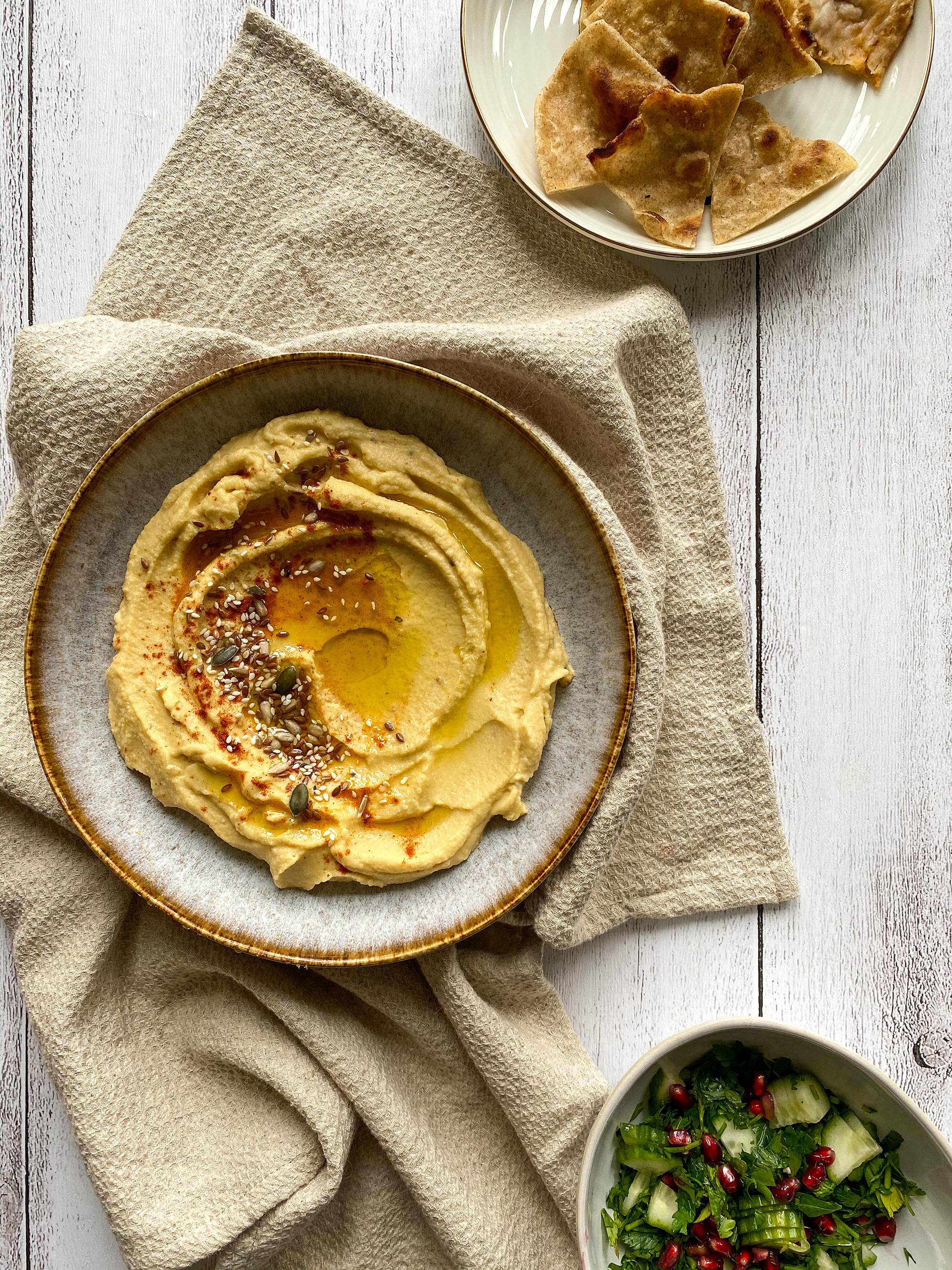 Light gray bowl with hummus garnished with some sesame seeds, flaxseeds and pumpkin seeds, olive oil. Side dishes are Tabbouleh and flatbreads. 