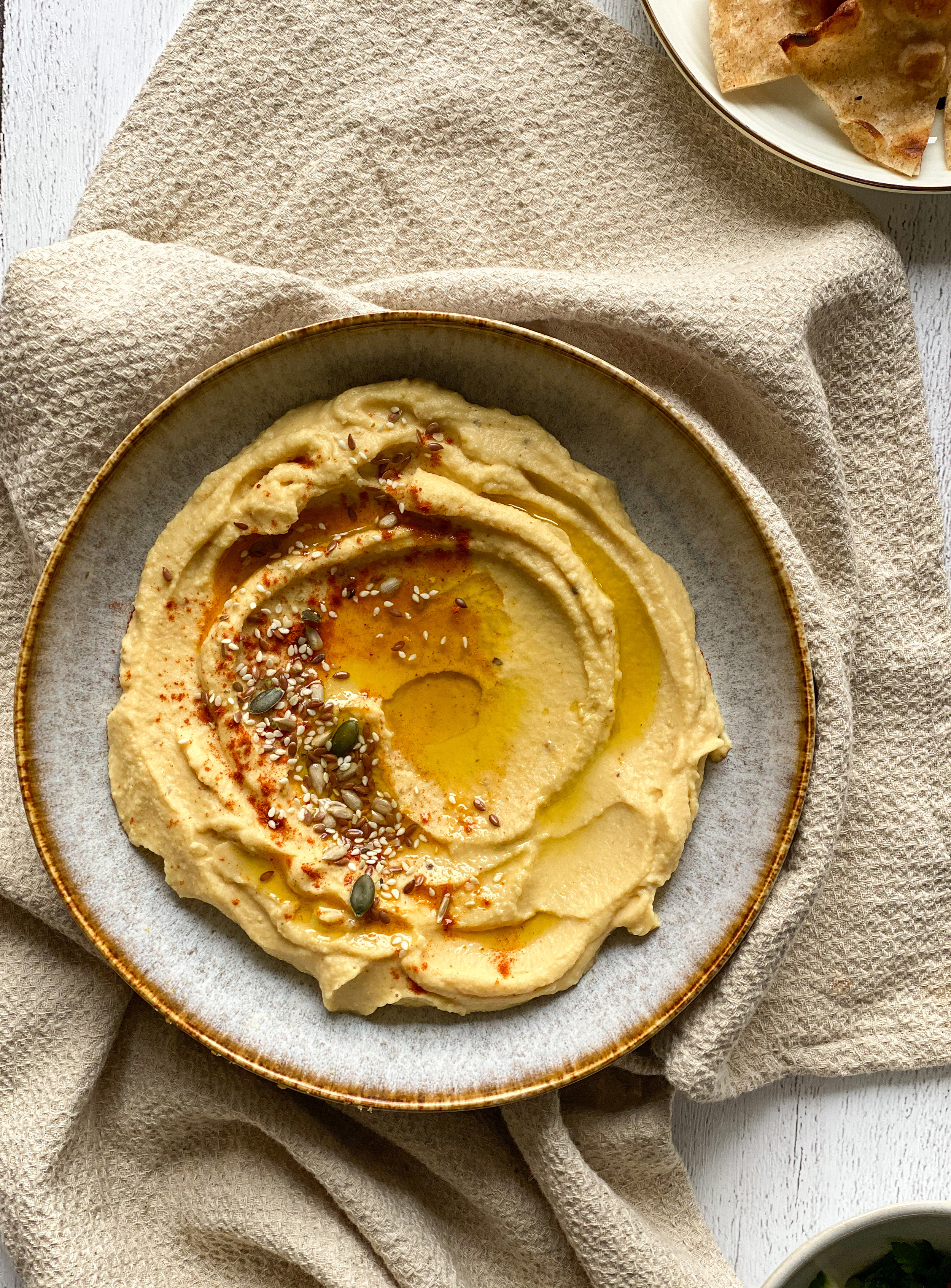 Light gray bowl with hummus garnished with some sesame seeds, flaxseeds and pumpkin seeds, olive oil. Side dishes are Tabbouleh and flatbreads. 